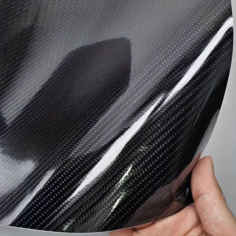A close-up of a glossy black carbon fiber textured film being held by a hand, showing the reflective surface and woven pattern.